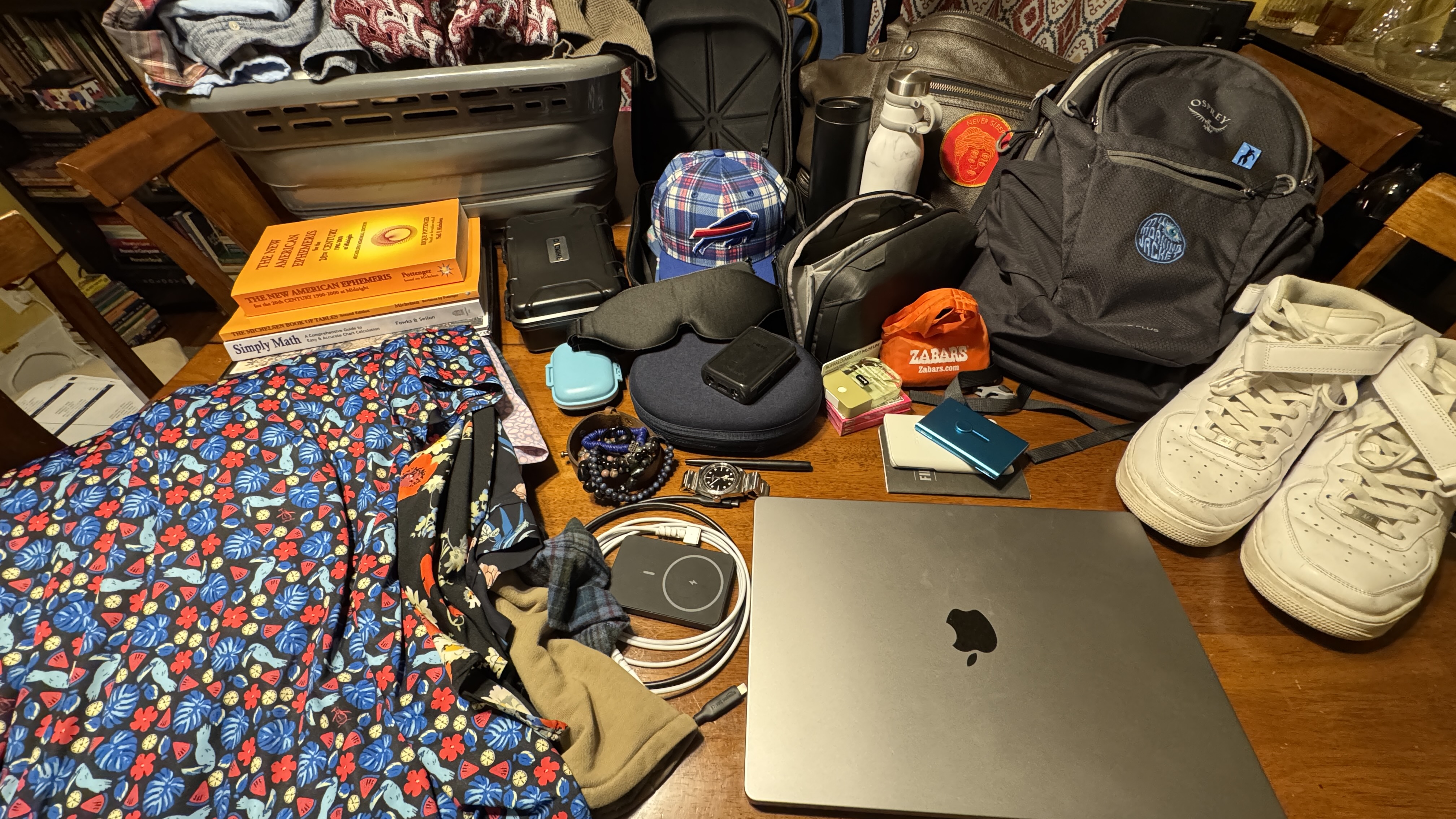 UAC 2026 test-packing spread on a dining room table: Osprey backpack, white Air Force 1s, MacBook Pro, ephemeris, DJI camera case, Buffalo Bills hat, and Hawaiian shirts